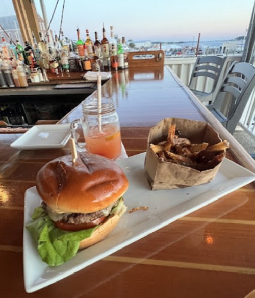 A burger with lettuce and tomato, served with fries and a drink, on a table with a bar and waterfront view in the background.