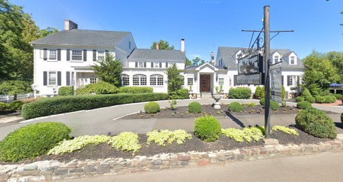 A large white house with a gray roof, surrounded by manicured lawns and shrubs, under a clear blue sky.