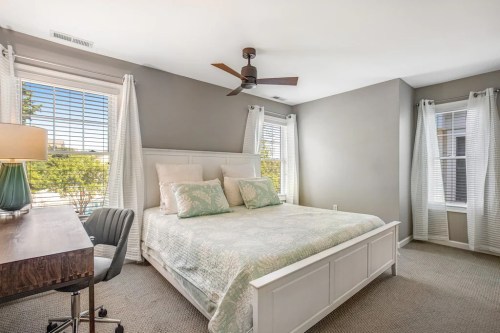 Bright bedroom with a queen bed, desk, and large windows, featuring light gray walls and soft green accents.