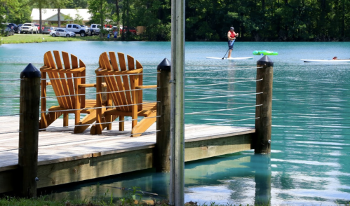 Two wooden chairs on a dock by a turquoise lake, with a person paddleboarding in the background.