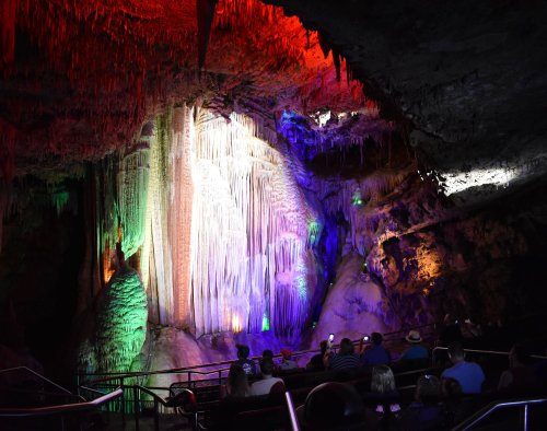Colorful illuminated stalactites and stalagmites in a cave, with visitors seated in the foreground.