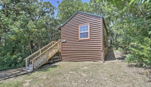 A brown log cabin with a staircase, surrounded by trees and greenery on a sunny day.