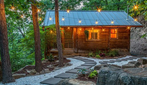 A cozy wooden cabin surrounded by trees, with string lights and a stone pathway leading to the entrance.