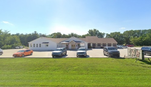 A building with a white exterior and a brown roof, surrounded by parked cars and green grass under a clear blue sky.