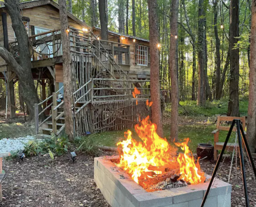 A cozy outdoor scene featuring a fire pit with flames, surrounded by trees and a treehouse in the background.