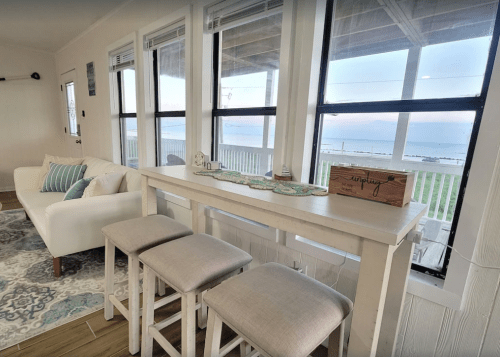 Cozy interior with a white sofa, bar stools, and large windows showcasing a view of the beach and ocean.