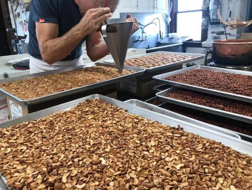 A person pipes caramel onto trays of roasted nuts in a kitchen, with various nut trays in the foreground and background.