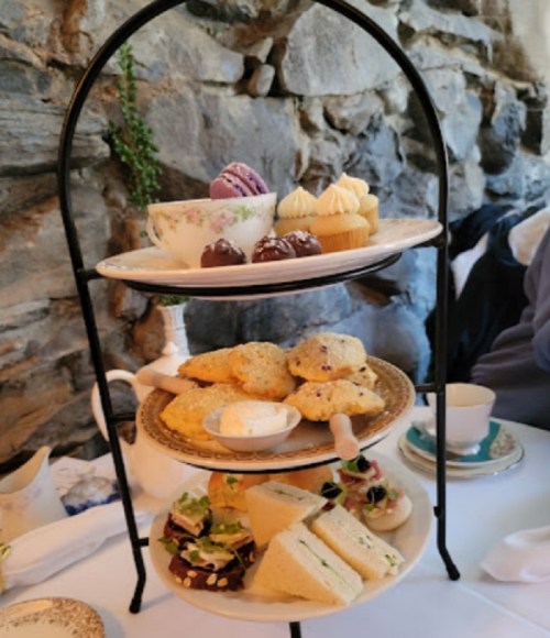 A tiered stand with assorted pastries, sandwiches, and a cup of tea, set against a stone wall backdrop.