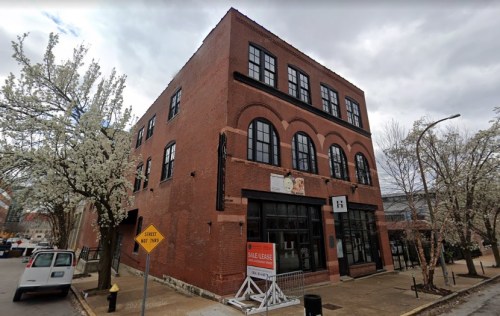 Red brick building with large windows, surrounded by flowering trees, featuring a "For Sale" sign in front.