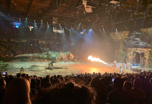 A large dinosaur on stage breathes fire in front of a cheering crowd at a live performance.