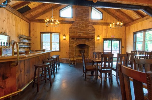 Cozy interior of a rustic bar with wooden walls, a stone fireplace, and seating around a central bar area.