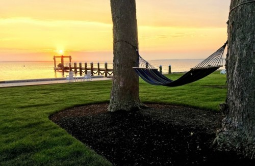 A hammock between two trees at sunset, overlooking a calm waterway and a wooden dock.