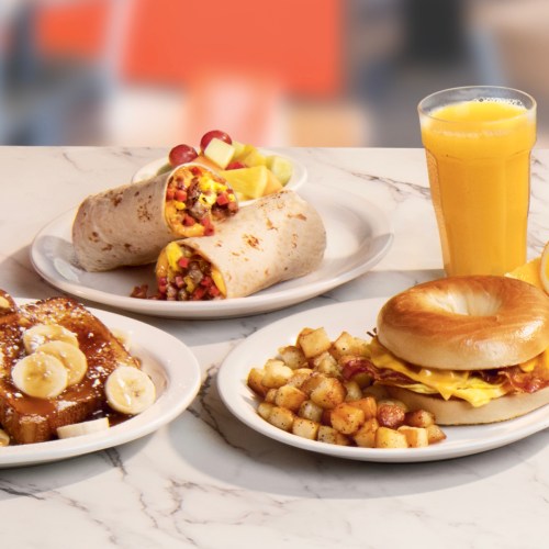 A table with a breakfast spread: two wraps with fruit, a bagel sandwich, and a plate of French toast with bananas.