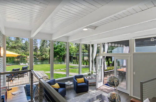 Bright, modern living space with large windows overlooking a green yard and water, featuring blue chairs and a sleek design.