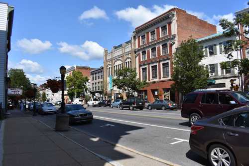 A street view featuring historic buildings, parked cars, and trees under a blue sky with scattered clouds.