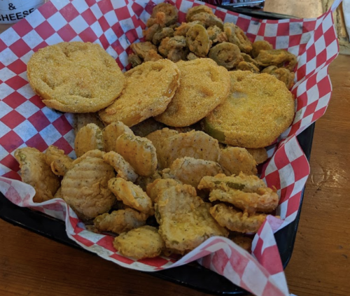 A basket of fried snacks, including golden fried green tomatoes and crispy pickles, served on red and white checkered paper.