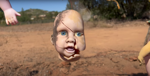 A cracked doll head with blue eyes hangs in a barren landscape, surrounded by dry earth and sparse vegetation.
