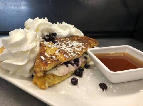 A plate with a stuffed French toast topped with whipped cream, blueberries, and a side of syrup.