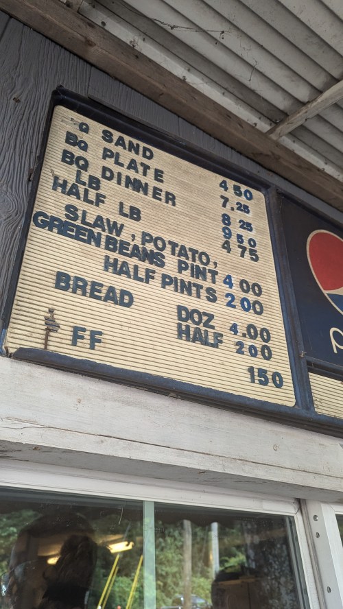 Menu board displaying prices for BBQ sandwiches, plates, dinners, sides, and bread. Prices range from $1.50 to $9.28.