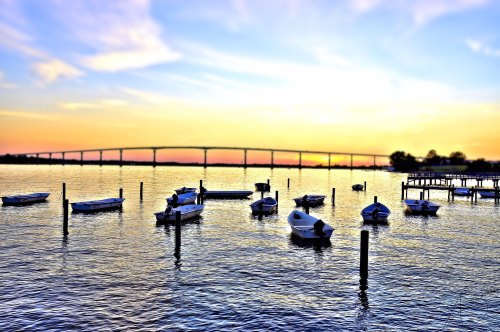 Boats anchored in calm water at sunset, with a bridge silhouetted against a colorful sky.