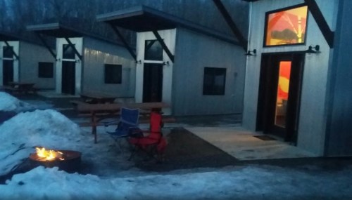 A cozy outdoor scene with modern cabins, a fire pit, and chairs, surrounded by snow and trees at dusk.