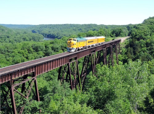 A yellow and green train crosses a tall, rust-colored bridge over a lush green landscape and river below.