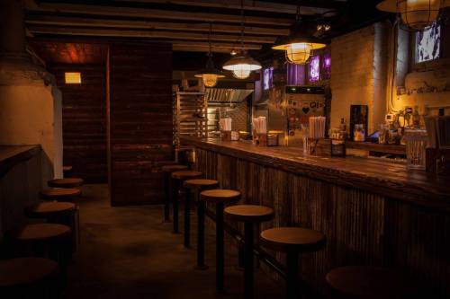 A dimly lit bar with wooden stools and a rustic counter, featuring vintage lighting and exposed brick walls.