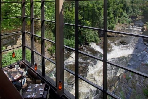 A modern restaurant with large windows overlooking a flowing river and lush greenery.