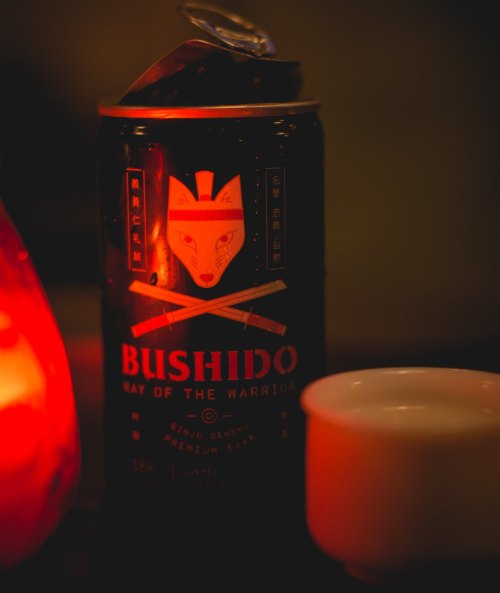 A can of Bushido sake featuring a fox design and crossed swords, next to a small white cup and a glowing red lamp.