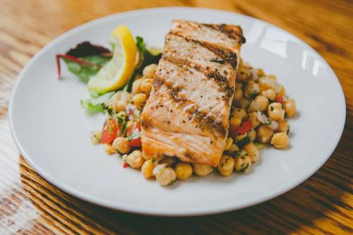 Grilled salmon fillet on a bed of chickpeas and vegetables, garnished with a lemon wedge and mixed greens.
