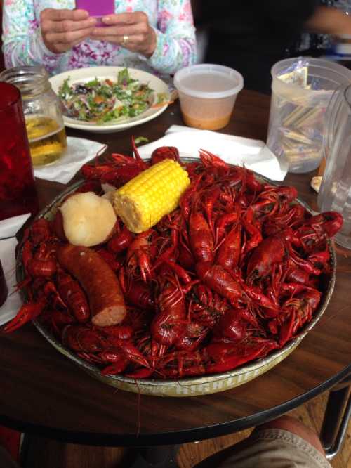 A large platter of boiled crawfish, sausage, corn, and a roll, with drinks and a salad in the background.