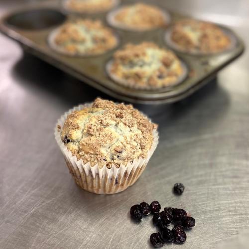 A freshly baked muffin with a crumbly topping, placed beside dried berries, with more muffins in a tray in the background.