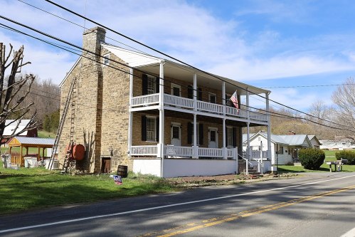 A two-story stone house with a porch, surrounded by greenery and power lines, along a rural road.