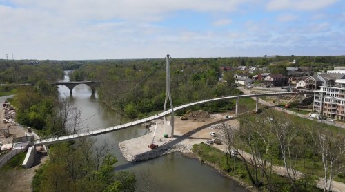 Aerial view of a suspension bridge over a river, surrounded by greenery and buildings in the background.