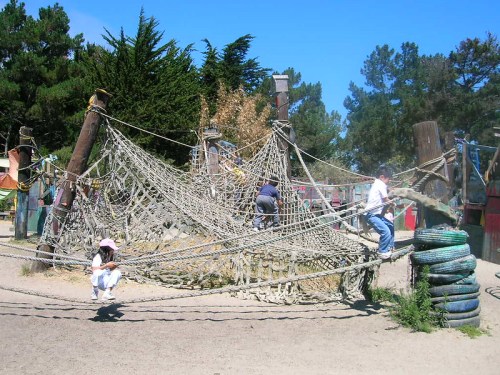 Children play on a large rope climbing structure in a sunny playground surrounded by trees.