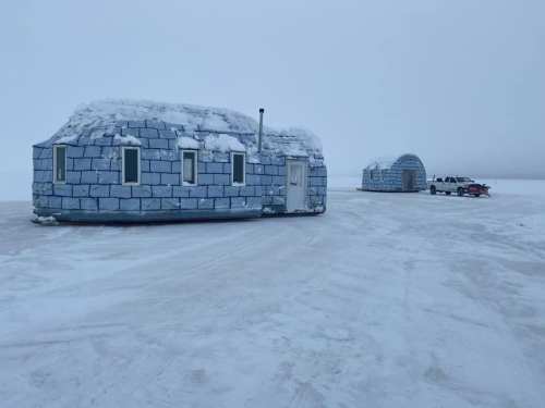 Two igloo-like structures made of ice blocks sit on a snowy landscape, with a vehicle parked nearby.