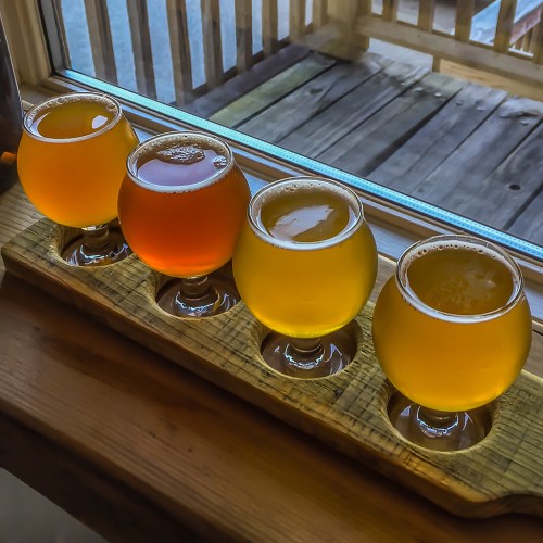 A wooden flight board holds four different craft beers, displayed in round glasses, with a scenic window view behind.