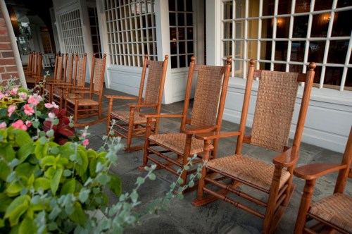 A row of wooden rocking chairs lined up on a porch, with colorful flowers in the foreground.