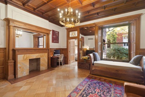 Cozy room with wooden paneling, a fireplace, a desk, and a window seat overlooking a tree-lined street.