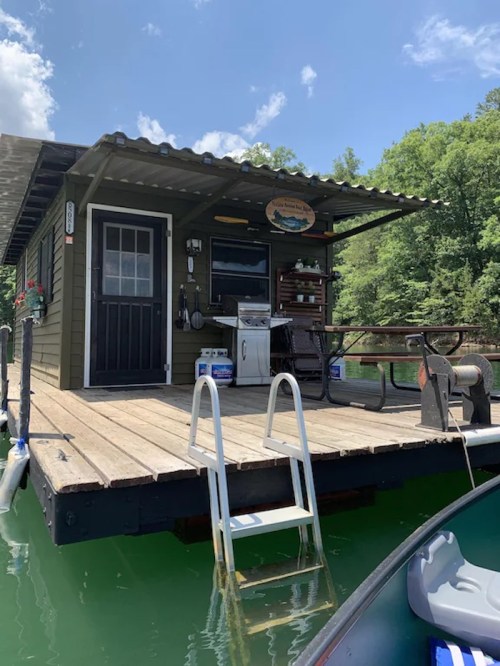 A cozy houseboat with a porch, stairs, and outdoor seating, surrounded by green trees and calm water.