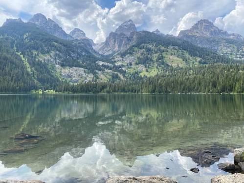 A serene lake reflecting mountains and clouds, surrounded by lush greenery and pine trees.