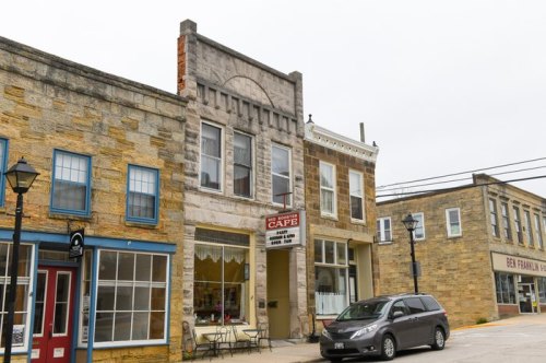 Historic stone buildings line a street, featuring a café and a parked car in a quaint town setting.
