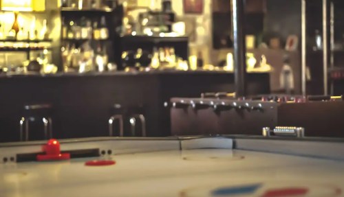 A close-up of an air hockey table in a dimly lit bar, with blurred background elements and a red puck on the surface.