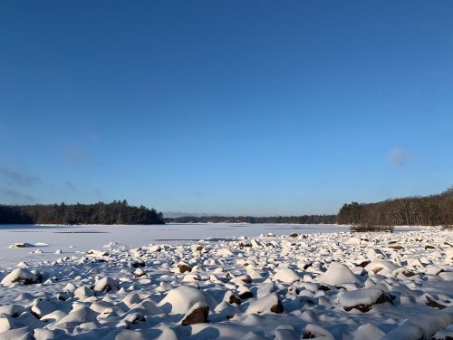 A snowy landscape with large rocks by a frozen lake under a clear blue sky and distant trees.