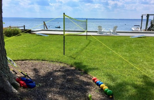 A grassy area by the water with a volleyball net, colorful balls, and chairs in the background under a blue sky.