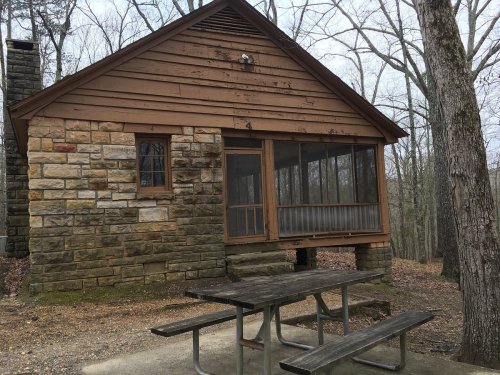 A rustic cabin with a stone and wood exterior, featuring a screened porch and a picnic table in a wooded area.