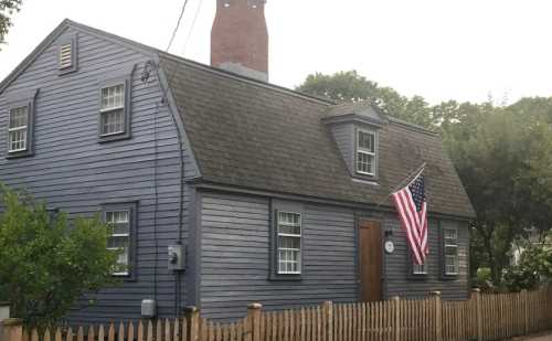 A blue house with a chimney and an American flag, surrounded by a wooden fence and greenery.