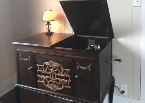 A vintage wooden cabinet with an open lid, revealing a turntable, next to a small lamp.