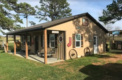 A rustic cabin with a porch, surrounded by grass and trees, featuring vintage signs and decorative wheels.