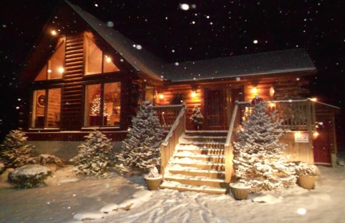 A cozy log cabin illuminated at night, surrounded by snow-covered trees and a snowy pathway. Snowflakes are falling.